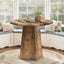 Wooden dining table with a round top and a single pedestal base in a room with large windows.
